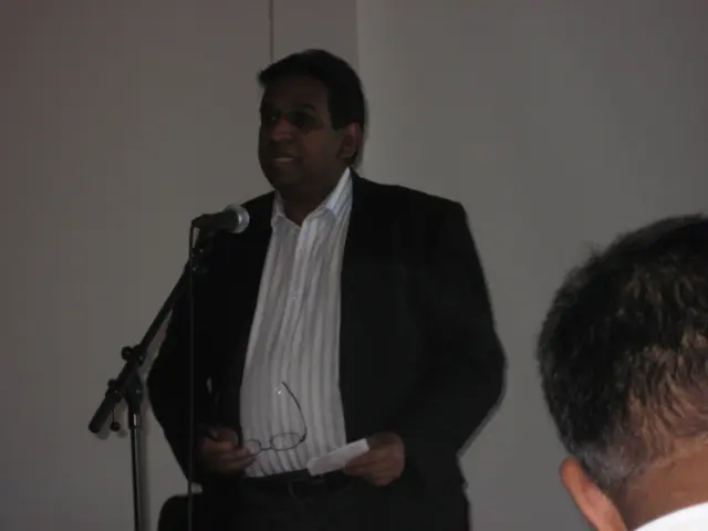 In this image there is a person speaking in the microphone holding paper in one hand and spectacles...