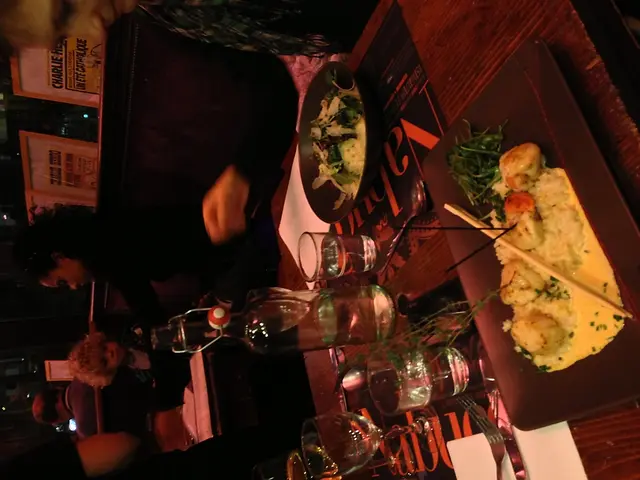 In this image we can a bottle, glasses, table, food items, plates, chopsticks, fork, spoons,...