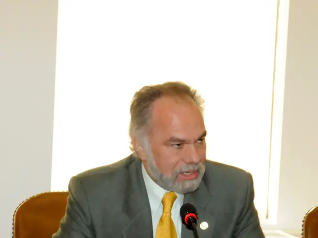 There is a man sitting in front of the table and speaking in a micro phone. behind him here is a...